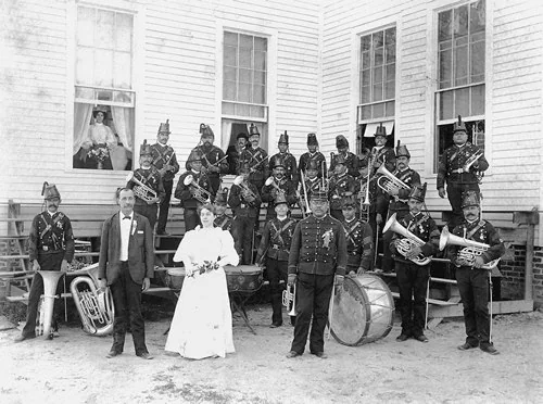 The Mexican Fourth Cavalry Band in dark uniforms and plumed helmets posed with brass instruments and a bass drum in front of a white frame building, with a woman in a white dress at the center