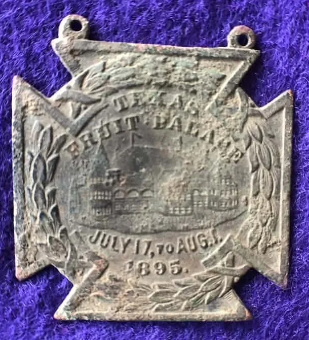 Cross-shaped 1895 souvenir medal with two hanging loops and laurel branches, showing the Texas Fruit Palace building and the words Texas Fruit Palace, July 17 to Aug. 1, 1895