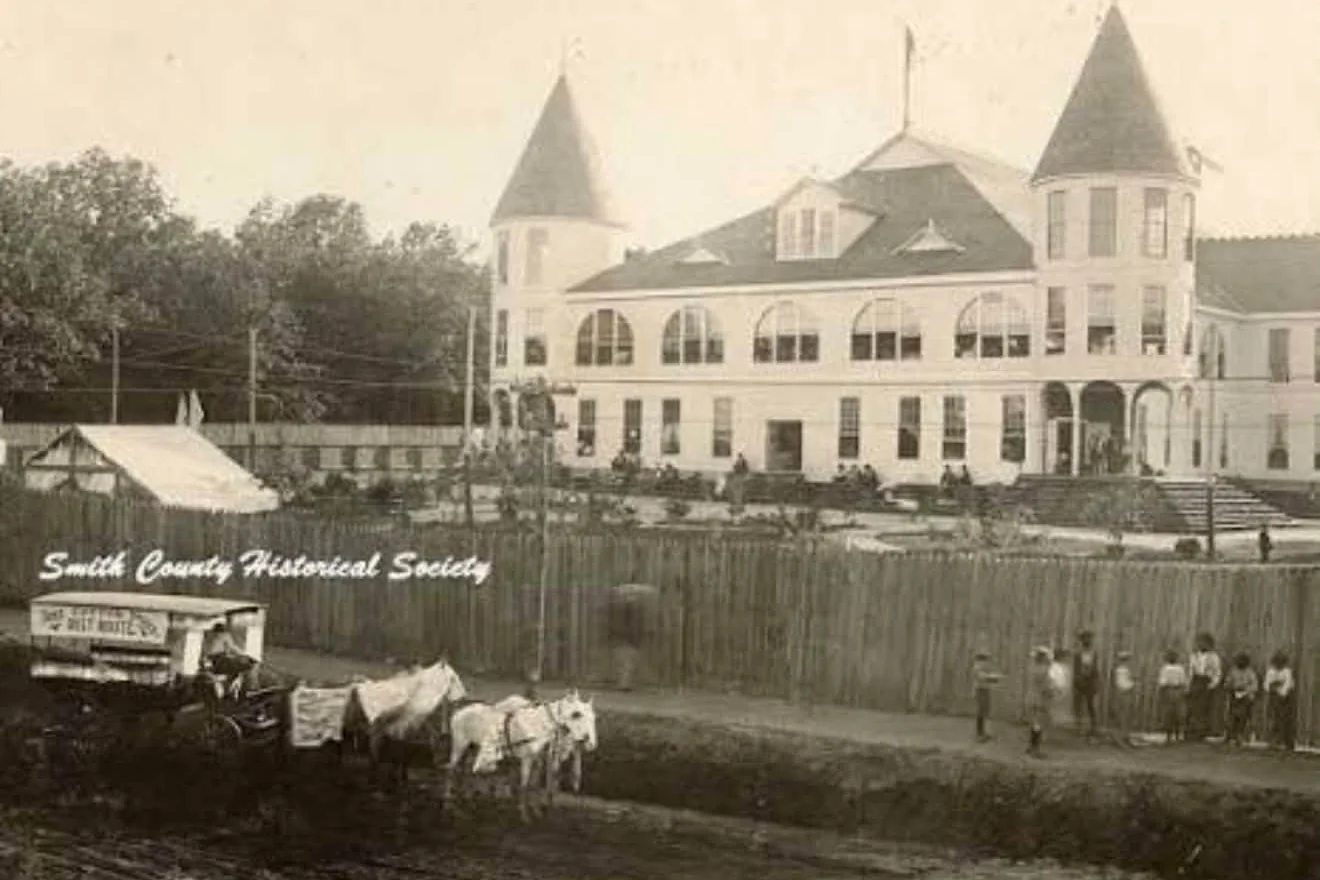 Photograph of the Texas Fruit Palace in Tyler: a white two-story frame building with two conical towers, a row of arched upper windows and a flagpole, behind a tall board fence, with a horse-drawn delivery wagon and a group of boys on the dirt street in front