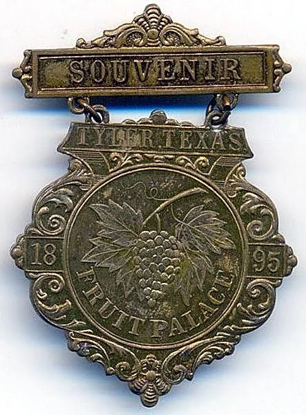Brass 1895 souvenir medal hanging from a bar reading Souvenir, stamped Tyler Texas, 1895, Fruit Palace, with a cluster of grapes and leaves in the center